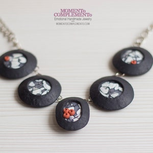 以下が含まれることがあります： 黒色のネックレスに、丸いペンダントが5つ付いています。各ペンダントには、白と黒のパターンが描かれたデザインが施されています。中央のペンダントには、赤いビーズが小さな塊になっています。