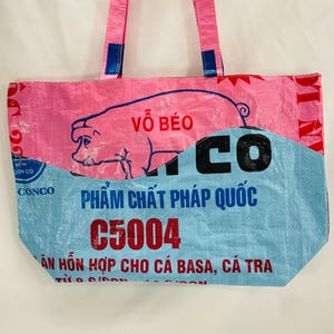 May include: Pink and blue tote bag made from recycled materials. The bag features a pig illustration with the text "V&Otilde; B&Eacute;O" and "PHẨM CHẤT PH&Aacute;P QUỐC C5004" printed on it.