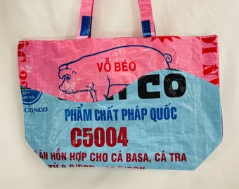 May include: Pink and blue tote bag made from recycled materials. The bag features a pig illustration with the text "VÕ BÉO" and "PHẨM CHẤT PHÁP QUỐC C5004" printed on it.