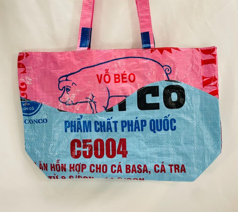May include: Pink and blue tote bag made from recycled materials. The bag features a pig illustration with the text "V&Otilde; B&Eacute;O" and "PHẨM CHẤT PH&Aacute;P QUỐC C5004" printed on it.