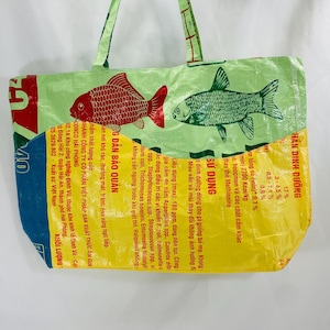 May include: A green and yellow reusable tote bag made from recycled plastic bags. The bag features a red and white fish and a green and white fish. The bag has a yellow section with text in Vietnamese that reads "THẦN DINH DƯỠNG" and "SỬ DỤNG".