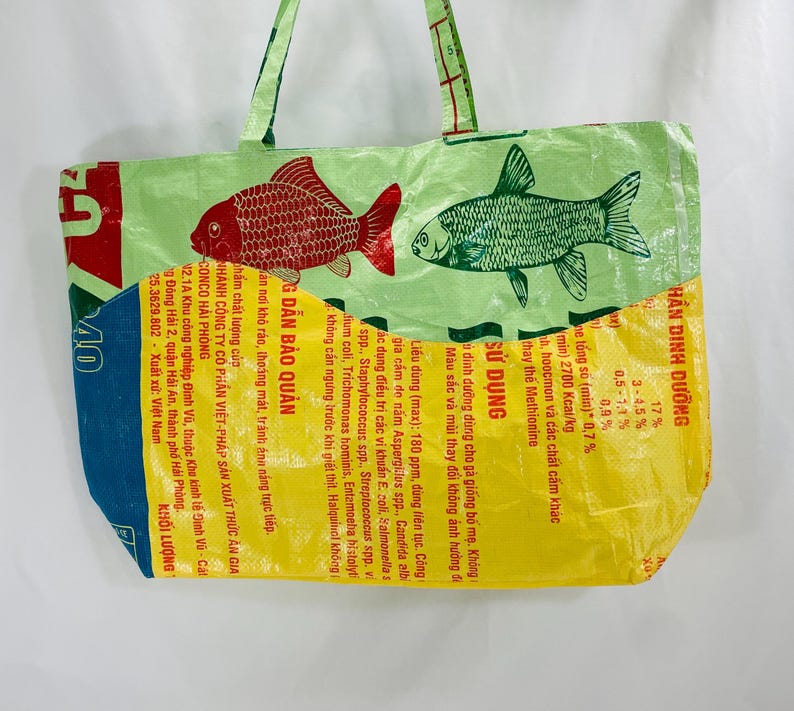 May include: A green and yellow reusable tote bag made from recycled plastic bags. The bag features a red and white fish and a green and white fish. The bag has a yellow section with text in Vietnamese that reads "THẦN DINH DƯỠNG" and "SỬ DỤNG".