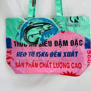 May include: A pink and green reusable tote bag made from recycled plastic bags. The bag features a graphic of a fish and the text "THỰC ĂN SI&Ecirc;U ĐẬM ĐẶC HEO TỪ 15KG ĐẾN XUẤT SẢN PHẨM CHẤT LƯỢNG CAO".