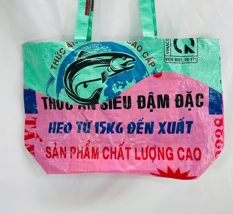 May include: A pink and green reusable tote bag made from recycled plastic bags. The bag features a graphic of a fish and the text "THỰC ĂN SI&Ecirc;U ĐẬM ĐẶC HEO TỪ 15KG ĐẾN XUẤT SẢN PHẨM CHẤT LƯỢNG CAO".