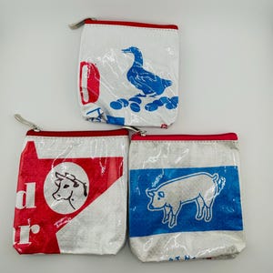 May include: Three square zippered pouches made from recycled woven plastic material. One pouch features a blue duck graphic, another displays a red and white cow illustration with partial text, and the third shows a white pig on a blue background.