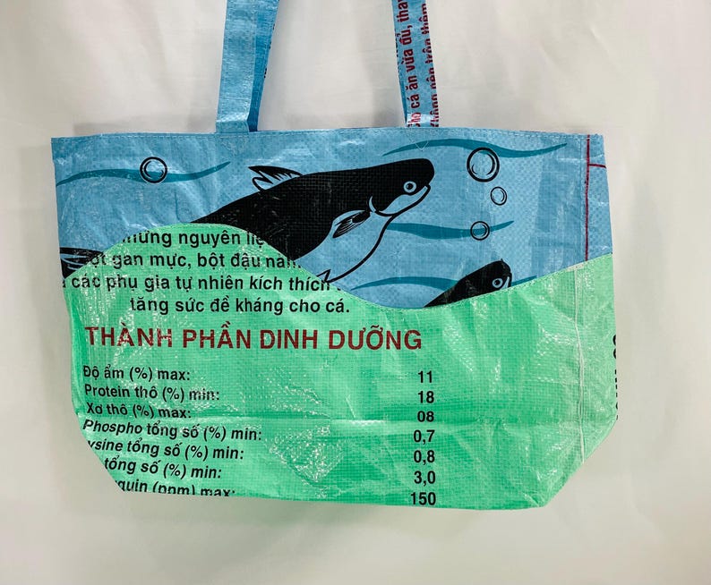May include: A blue and green reusable tote bag with a fish graphic and the text "TH&Agrave;NH PHẦN DINH DƯỠNG" and a list of nutritional information. The bag is made from recycled plastic.