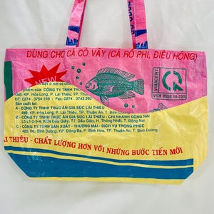 May include: A pink and yellow reusable tote bag with a green border. The bag has a fish design and the text "D&Ugrave;NG CHO C&Aacute; C&Oacute; V&Acirc;Y (C&Aacute; R&Ocirc; PHI, ĐI&Ecirc;U HỒNG)" and "VINACERT" printed on it.