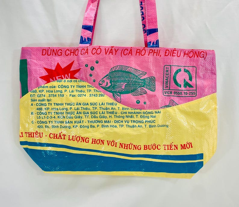 May include: A pink and yellow reusable tote bag with a green border. The bag has a fish design and the text "D&Ugrave;NG CHO C&Aacute; C&Oacute; V&Acirc;Y (C&Aacute; R&Ocirc; PHI, ĐI&Ecirc;U HỒNG)" and "VINACERT" printed on it.