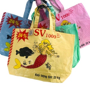 May include: Three reusable shopping bags with a mermaid design. The bags are yellow, blue, and pink with black text that reads "SV 1000" and "24%". The yellow bag has a mermaid with red hair and a red tail. The text on the yellow bag also reads "Khối lượng tịnh: 25 kg".