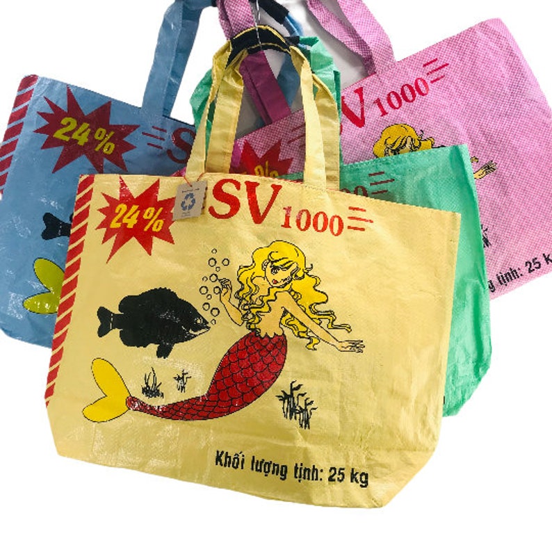 May include: Three reusable shopping bags with a mermaid design. The bags are yellow, blue, and pink with black text that reads "SV 1000" and "24%". The yellow bag has a mermaid with red hair and a red tail. The text on the yellow bag also reads "Khối lượng tịnh: 25 kg".