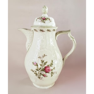 May include: Cream-colored porcelain coffee pot with a lid, featuring a floral rose design in pink and green. The pot has a curved spout, a decorative handle, and embossed detailing. A classic piece for serving coffee or tea.