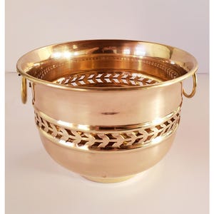 May include: A polished brass bowl with a decorative cut-out band around the middle. The bowl has two small handles and a perforated interior base. The overall design is a classic, elegant style.