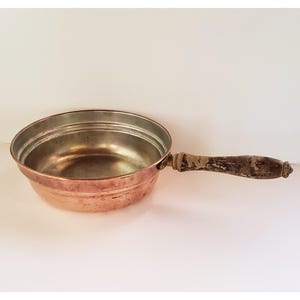 Op de afbeelding: Een koperen steelpan met een geklonken handvat. De pan heeft een afgeronde komvorm met een zilverkleurig interieur en een koperen buitenkant. De handgreep is van hout en vertoont gebruikssporen.