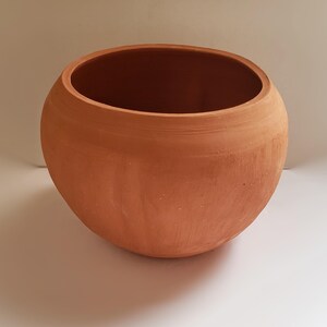 Op de afbeelding: Een ronde, terracotta-kleurige kleipot met een brede, open bovenkant en een gladde, matte afwerking. De pot heeft een eenvoudige, bolvormige vorm met een licht taps toelopende rand, die de natuurlijke, aardse oranjebruine tint van het materiaal toont.
