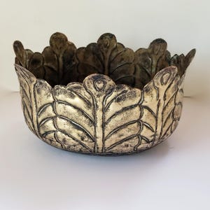 Puede incluir: Un cuenco redondo de metal con un acabado dorado desgastado. El exterior presenta un patrón de hojas en relieve que se repite hasta el borde festoneado, creando una silueta de corona. La superficie tiene acentos oscuros y envejecidos.