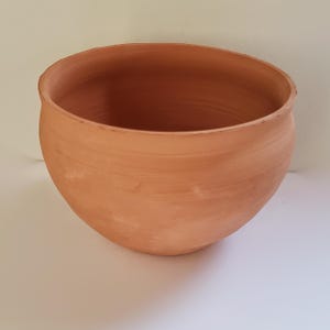 Puede incluir: Un cuenco de arcilla color terracota con una base profunda y redondeada y un acabado mate suave. El cuenco presenta estrías horizontales visibles en la superficie exterior, lo que indica una construcción artesanal con un borde circular abierto.