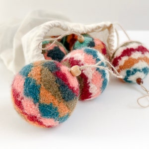 May include: A set of five colorful felt ornaments with geometric patterns in shades of red, pink, orange, green, blue, and brown. The ornaments are hanging from a white drawstring bag.