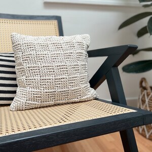 May include: A beige knitted throw pillow with a woven pattern sits on a black and wicker chair.
