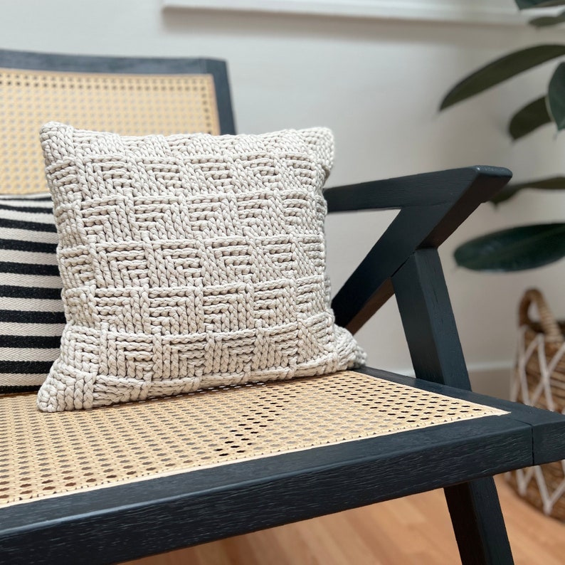 May include: A beige knitted throw pillow with a woven pattern sits on a black and wicker chair.