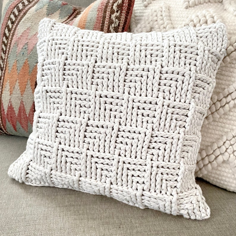 May include: A white knitted throw pillow with a woven pattern. The pillow is made of thick, chunky yarn and has a soft, textured surface.