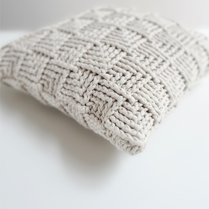 May include: A close-up of a cream-colored knitted throw pillow with a textured woven pattern.