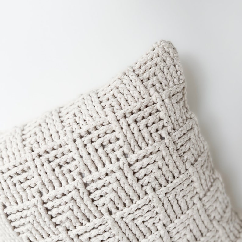 May include: A close-up of a cream-colored knitted pillow with a woven pattern.