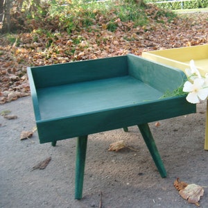 Op de afbeelding: Twee houten trays met poten, een groen geverfd en de andere geel, staan op een bed van gevallen bladeren. De groene tray heeft een witte bloem erop.