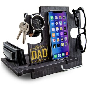 May include: A dark gray wooden desk organizer with slots for a phone, glasses, watch, keys, and wallet. The organizer has the text "Best Dad Ever" in gold lettering. A black watch with a leather band is displayed.