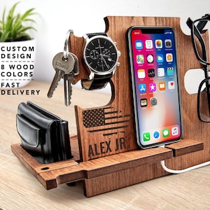 Puede incluir: Soporte de teléfono de madera con un nombre personalizado "ALEX JR." y un diseño de bandera estadounidense. El soporte también tiene ranuras para un reloj, llaves y gafas.