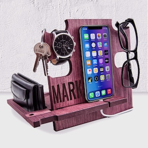 Può includere: Un organizer da scrivania in legno di colore rosso-viola scuro, che contiene uno smartphone, occhiali, un orologio, chiavi e un portafoglio. La parola "MARK" è incisa sulla parte anteriore. Il telefono è in carica con un cavo bianco.