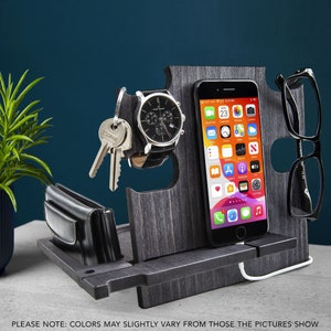 May include: A gray wooden desk organizer with slots for a phone, watch, keys, wallet, and glasses. The phone displays the date as Wednesday, the 23rd. The text at the bottom reads "PLEASE NOTE: COLORS MAY SLIGHTLY VARY FROM THOSE THE PICTURES SHOW."