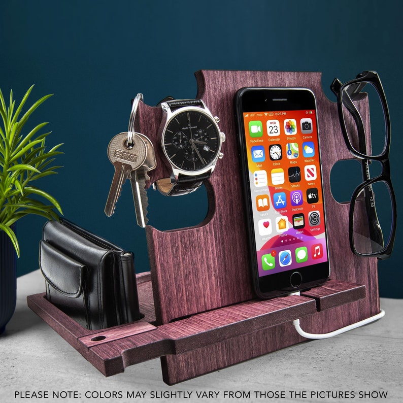 May include: A wooden desk organizer in a dark reddish-brown hue, holding a black watch, keys, a wallet, a smartphone, and eyeglasses. The smartphone displays app icons. The organizer has a charging cable and slots for various items. The text at the bottom says "PLEASE NOTE: COLORS MAY SLIGHTLY VARY FROM THOSE THE PICTURES SHOW."