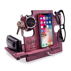 May include: A wooden desk organizer in a dark reddish-purple hue. It holds a smartphone, eyeglasses, a watch, keys, and a wallet. The organizer has the text "Hubby's Stuff William" engraved on it. A white charging cable is connected to the phone.
