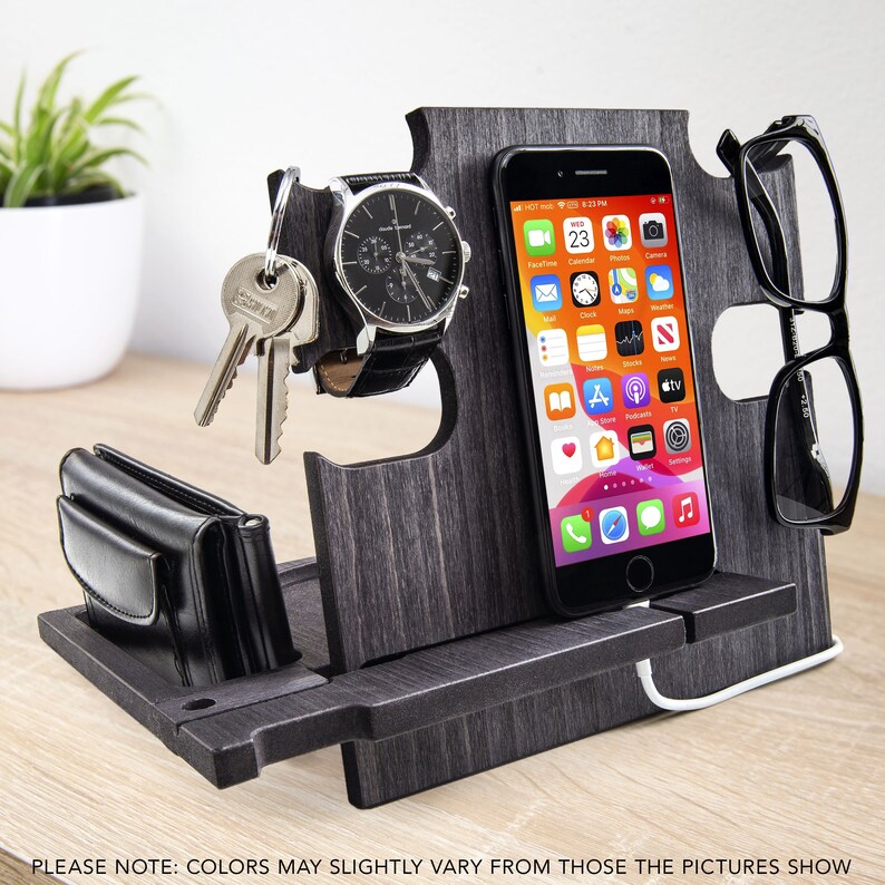 May include: A wooden desk organizer in a dark gray finish, holding a smartphone, watch, keys, wallet, and eyeglasses. The organizer has slots for each item and a charging cable. The text at the bottom says "COLORS MAY SLIGHTLY VARY FROM THOSE THE PICTURES SHOW."