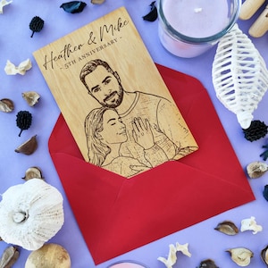 Può includere: Un biglietto di anniversario in legno con un ritratto inciso al laser di una coppia, in una busta rossa. Il biglietto recita "Heather & Mike - 5th Anniversary". Lo sfondo è viola con elementi decorativi.