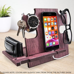 May include: A wooden desk organizer in a dark reddish-brown finish. It holds a smartphone, eyeglasses, a watch, keys, and a wallet. The phone displays app icons. The organizer has a charging cable and slots for items. The text at the bottom says "PLEASE NOTE: COLORS MAY SLIGHTLY VARY FROM THOSE THE PICTURES SHOW."