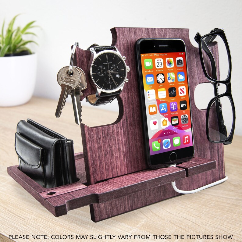 May include: A wooden desk organizer in a dark reddish-brown finish. It holds a smartphone, eyeglasses, a watch, keys, and a wallet. The phone displays app icons. The organizer has a charging cable and slots for items. The text at the bottom says "PLEASE NOTE: COLORS MAY SLIGHTLY VARY FROM THOSE THE PICTURES SHOW."