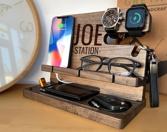 May include: A wooden phone charging station with a personalized name "JOE STATION" engraved on the front. It features slots for a phone, watch, glasses, and keys. The station is made of dark brown wood and has a sleek, modern design.