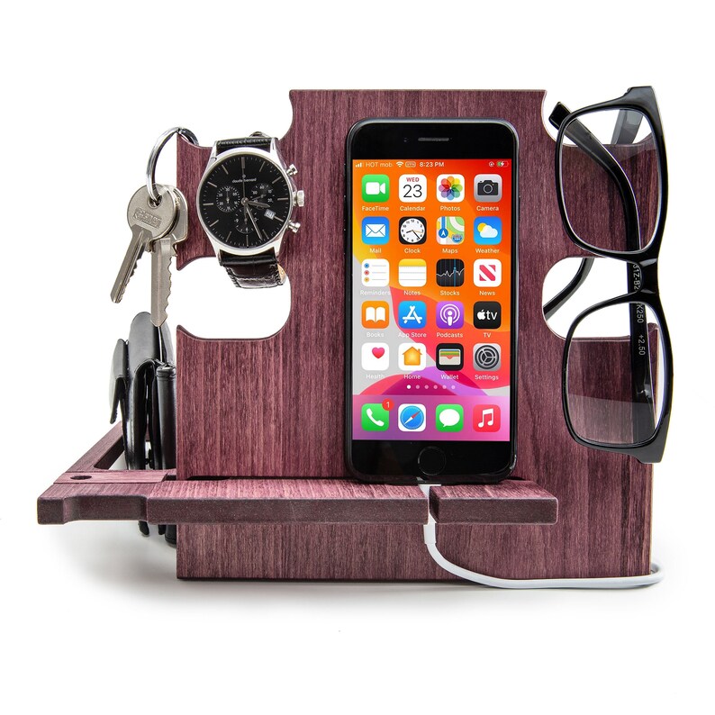 May include: A wooden desk organizer in a dark reddish-brown color. It holds a smartphone, eyeglasses, a watch, keys, and a wallet. The phone displays app icons. The watch has a black leather band. The eyeglasses are black.