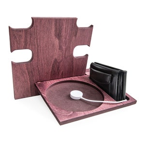 May include: A wooden charging station in a puzzle piece design, stained in a deep reddish-brown. It features a circular charging pad with a white Apple Watch charger, a black leather wallet, and a vertical back piece.