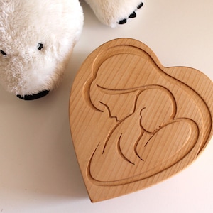 Peut inclure: Une bo&icirc;te en bois en forme de c&oelig;ur avec un motif sculpt&eacute; d'une m&egrave;re et d'un enfant. La bo&icirc;te est en bois de couleur claire et a une finition lisse. Un ours polaire en peluche blanc est en arri&egrave;re-plan.