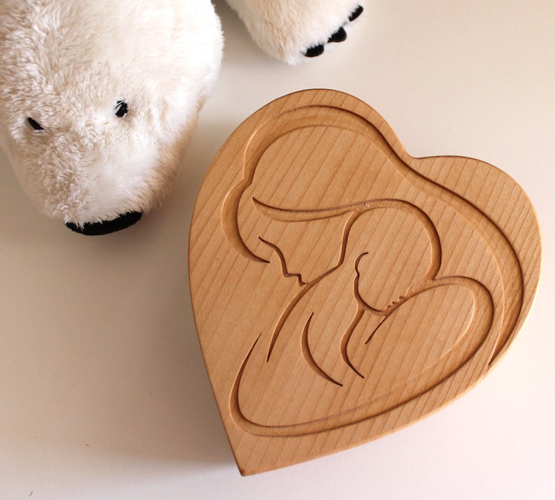 Peut inclure: Une bo&icirc;te en bois en forme de c&oelig;ur avec un motif sculpt&eacute; d'une m&egrave;re et d'un enfant. La bo&icirc;te est en bois de couleur claire et a une finition lisse. Un ours polaire en peluche blanc est en arri&egrave;re-plan.