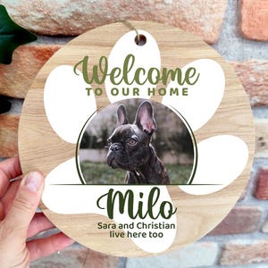 Può includere: Un cartello rotondo in legno con un design a impronta di zampa e la scritta "Welcome to our home". Al centro c'è una foto di un cane, con il nome "Milo" sotto. Il testo "Sara and Christian live here too" è in basso.