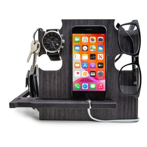 May include: A dark gray wooden desk organizer with slots for a smartphone, eyeglasses, watch, keys, and wallet. The smartphone displays a colorful app screen. A white charging cable is connected to the phone.