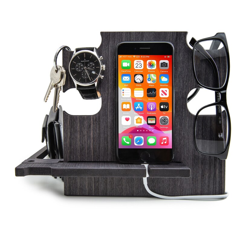 May include: A dark gray wooden desk organizer with slots for a smartphone, eyeglasses, watch, keys, and wallet. The smartphone displays a colorful app screen. A white charging cable is connected to the phone.
