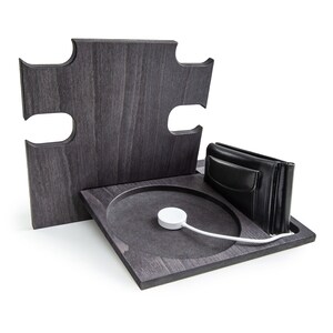 May include: A dark gray wooden charging station with a puzzle-piece design. It features a circular charging area with a white Apple Watch charger, a black leather wallet, and a vertical back piece.