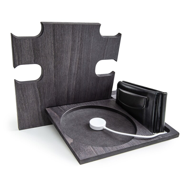 May include: A dark gray wooden charging station with a puzzle-piece design. It features a circular charging area with a white Apple Watch charger, a black leather wallet, and a vertical back piece.