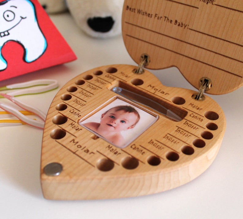 Peut inclure: Coffret en bois en forme de c&oelig;ur pour conserver les dents de b&eacute;b&eacute;. La bo&icirc;te est ouverte, r&eacute;v&eacute;lant une photo de b&eacute;b&eacute; et des compartiments &eacute;tiquet&eacute;s pour chaque dent. Le couvercle est grav&eacute; de "Best Wishes For The Baby". Un aimant ferme la bo&icirc;te. Une carte rouge avec une dent de dessin anim&eacute; est en arri&egrave;re-plan.