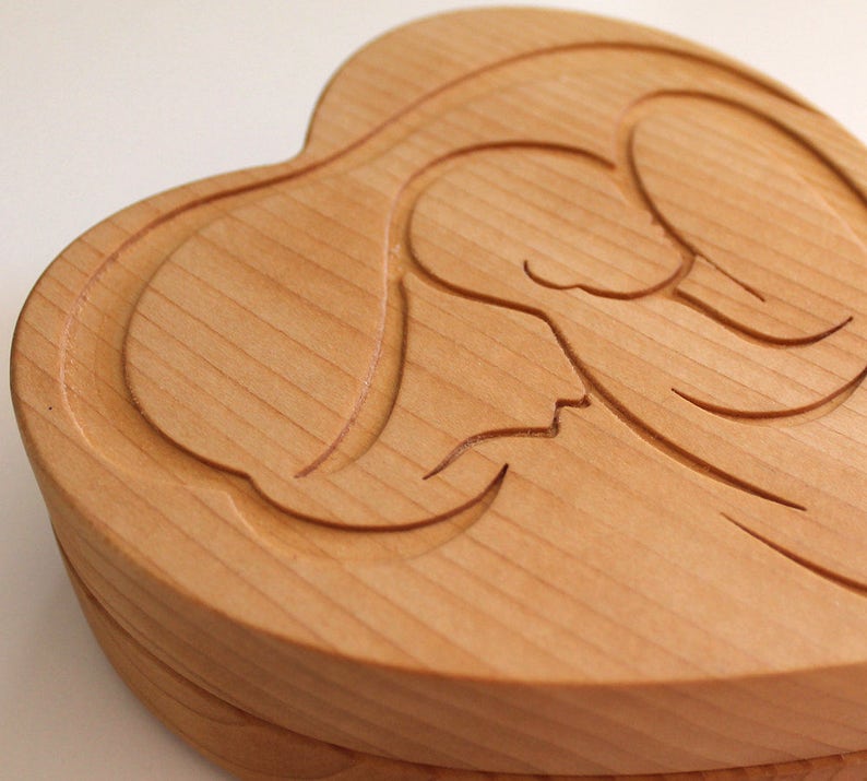 Peut inclure: Une bo&icirc;te en bois en forme de c&oelig;ur avec un motif d'ange sculpt&eacute; sur le couvercle. La bo&icirc;te est en bois de couleur claire, avec le grain du bois visible. Le motif de l'ange est sculpt&eacute; en relief, ajoutant de la profondeur et de la texture.