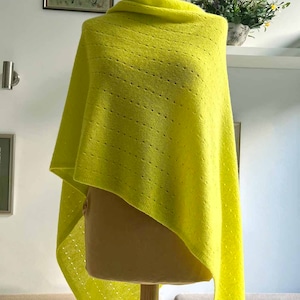 Op de afbeelding: Een felgele gebreide poncho met een delicaat opengewerkt patroon. De poncho is over een mannequin gedrapeerd en heeft een driehoekige vorm.